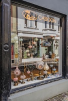 A shop window display featuring an array of ornate, colorful lamps with intricate mosaic designs. Various shapes and sizes of lamps are arranged on a narrow wooden shelf, creating an inviting and artistic presentation. The background shows a glimpse of the shop's interior with more decorative items and reflections of the street outside.