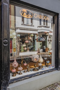A shop window display featuring an array of ornate, colorful lamps with intricate mosaic designs. Various shapes and sizes of lamps are arranged on a narrow wooden shelf, creating an inviting and artistic presentation. The background shows a glimpse of the shop's interior with more decorative items and reflections of the street outside.
