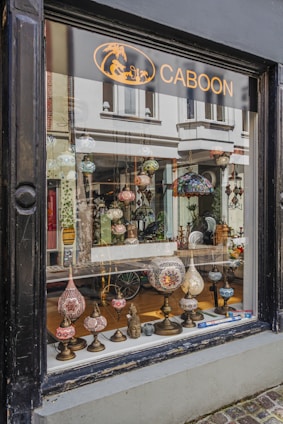 A shop window display featuring an array of ornate, colorful lamps with intricate mosaic designs. Various shapes and sizes of lamps are arranged on a narrow wooden shelf, creating an inviting and artistic presentation. The background shows a glimpse of the shop's interior with more decorative items and reflections of the street outside.