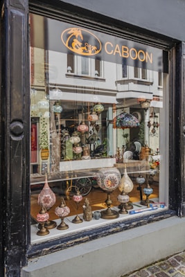 A shop window display featuring an array of ornate, colorful lamps with intricate mosaic designs. Various shapes and sizes of lamps are arranged on a narrow wooden shelf, creating an inviting and artistic presentation. The background shows a glimpse of the shop's interior with more decorative items and reflections of the street outside.