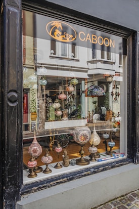 A shop window display featuring an array of ornate, colorful lamps with intricate mosaic designs. Various shapes and sizes of lamps are arranged on a narrow wooden shelf, creating an inviting and artistic presentation. The background shows a glimpse of the shop's interior with more decorative items and reflections of the street outside.