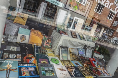 A storefront window display is filled with various books, primarily focused on art styles like Art Deco and Art Nouveau. The books are arranged in an eye-catching manner, with colorful covers featuring geometric and artistic designs. Reflections of the street, showing bicycles and buildings, blend with the display.
