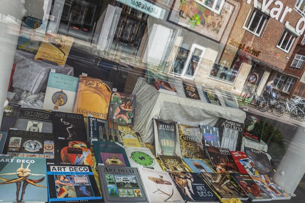 A storefront window display is filled with various books, primarily focused on art styles like Art Deco and Art Nouveau. The books are arranged in an eye-catching manner, with colorful covers featuring geometric and artistic designs. Reflections of the street, showing bicycles and buildings, blend with the display.