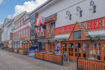 A street view featuring a series of colorful buildings with a European architectural style. The prominent establishment appears to be a pub or beer hall, with red letters spelling out 'Bier' and various flags, including Norwegian and Austrian, hanging above. Wooden accents and decorative lights add to its inviting, rustic charm.