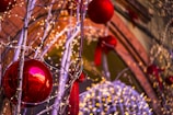 A festive decoration featuring large red baubles hanging from branches adorned with warm white string lights. The background includes a mix of additional lights and red ribbon decorations, creating a warm and festive atmosphere.