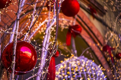 A festive decoration featuring large red baubles hanging from branches adorned with warm white string lights. The background includes a mix of additional lights and red ribbon decorations, creating a warm and festive atmosphere.