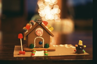 A gingerbread house decorated with colorful candies sits on a table. It features a peppermint candy on the front and gummy candies lining the roof. There's a candy path leading up to the door, and a small candy Christmas tree with a gumdrop at the top to the right. Bokeh lights in the background add a warm, festive feel.