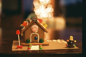A gingerbread house decorated with colorful candies sits on a table. It features a peppermint candy on the front and gummy candies lining the roof. There's a candy path leading up to the door, and a small candy Christmas tree with a gumdrop at the top to the right. Bokeh lights in the background add a warm, festive feel.