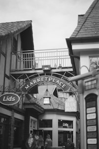 A monochrome photograph captures a small marketplace entrance with an arch labeled 'Marketplace.' The architecture features pitched roofs and wooden elements, giving a cozy, traditional feel. People walk through the area, with some entering the 'Lids' store. An adjacent sign reads 'Little Blue House.' The setting appears casual and inviting.