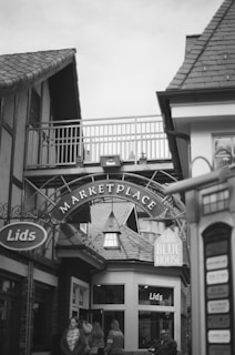 A monochrome photograph captures a small marketplace entrance with an arch labeled 'Marketplace.' The architecture features pitched roofs and wooden elements, giving a cozy, traditional feel. People walk through the area, with some entering the 'Lids' store. An adjacent sign reads 'Little Blue House.' The setting appears casual and inviting.