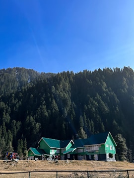 A scenic view of a green-roofed building nestled at the base of a dense forested mountain range under a clear blue sky. A group of people stand in front of the building, which is surrounded by tall coniferous trees.