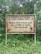 A weathered sign stands in front of a lush, green forest backdrop. The sign, bordered in red, bears a message in Indonesian about nature conservation and obeying divine commandments. The surrounding environment is dense with various shades of green foliage and trees.
