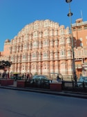 A large, intricately designed building featuring numerous windows and stone decorations, constructed with a pinkish hue, typical of traditional Indian architecture. The structure dominates the scene under a clear blue sky, with several people walking along the street and a few vehicles, including an auto rickshaw, visible in the foreground.