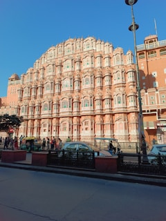A large, intricately designed building featuring numerous windows and stone decorations, constructed with a pinkish hue, typical of traditional Indian architecture. The structure dominates the scene under a clear blue sky, with several people walking along the street and a few vehicles, including an auto rickshaw, visible in the foreground.