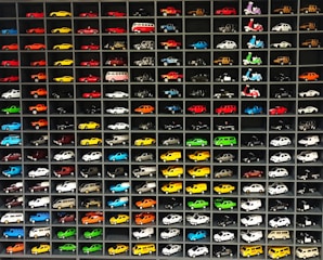 A collection of miniature toy vehicles arranged neatly in a grid of shelves. Various models of cars, vans, and scooters are displayed, featuring a wide range of vibrant colors.