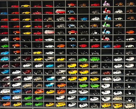 A collection of miniature toy vehicles arranged neatly in a grid of shelves. Various models of cars, vans, and scooters are displayed, featuring a wide range of vibrant colors.