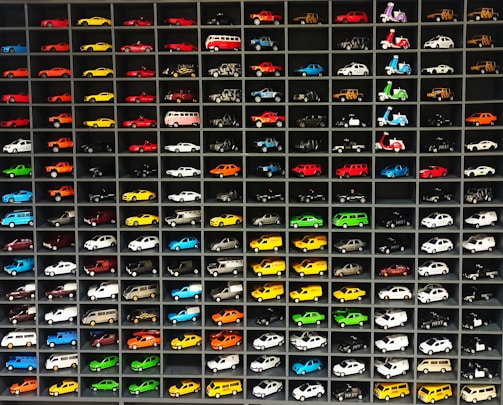 A collection of miniature toy vehicles arranged neatly in a grid of shelves. Various models of cars, vans, and scooters are displayed, featuring a wide range of vibrant colors.