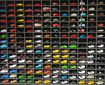 A collection of miniature toy vehicles arranged neatly in a grid of shelves. Various models of cars, vans, and scooters are displayed, featuring a wide range of vibrant colors.