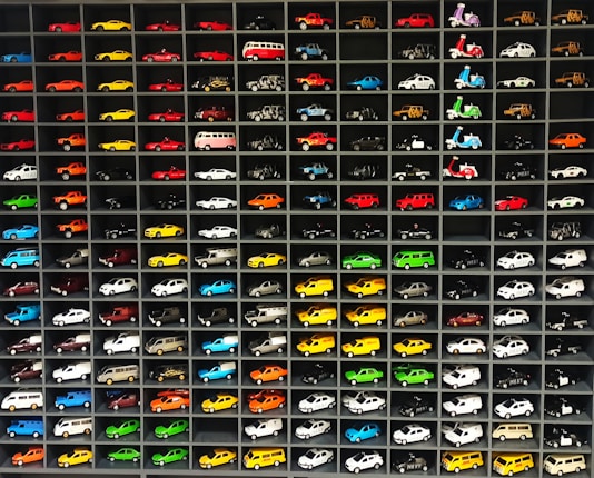 A collection of miniature toy vehicles arranged neatly in a grid of shelves. Various models of cars, vans, and scooters are displayed, featuring a wide range of vibrant colors.