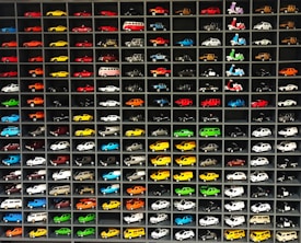 A collection of miniature toy vehicles arranged neatly in a grid of shelves. Various models of cars, vans, and scooters are displayed, featuring a wide range of vibrant colors.