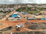 An aerial view captures a construction site with machinery and materials scattered across a reddish dirt ground. Several buildings and storage units are positioned at the site, along with a small number of vehicles. Surroundings include residential structures, green areas with palm trees, and hilly terrain in the background under a cloudy sky.