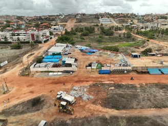 An aerial view captures a construction site with machinery and materials scattered across a reddish dirt ground. Several buildings and storage units are positioned at the site, along with a small number of vehicles. Surroundings include residential structures, green areas with palm trees, and hilly terrain in the background under a cloudy sky.