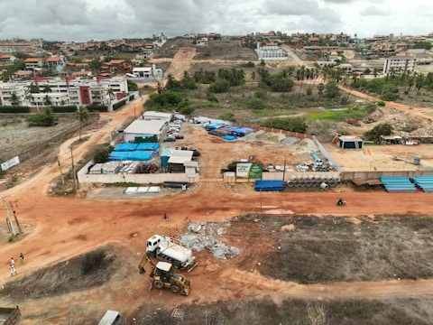 An aerial view captures a construction site with machinery and materials scattered across a reddish dirt ground. Several buildings and storage units are positioned at the site, along with a small number of vehicles. Surroundings include residential structures, green areas with palm trees, and hilly terrain in the background under a cloudy sky.