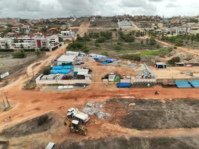 An aerial view captures a construction site with machinery and materials scattered across a reddish dirt ground. Several buildings and storage units are positioned at the site, along with a small number of vehicles. Surroundings include residential structures, green areas with palm trees, and hilly terrain in the background under a cloudy sky.