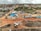 An aerial view captures a construction site with machinery and materials scattered across a reddish dirt ground. Several buildings and storage units are positioned at the site, along with a small number of vehicles. Surroundings include residential structures, green areas with palm trees, and hilly terrain in the background under a cloudy sky.