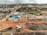 An aerial view captures a construction site with machinery and materials scattered across a reddish dirt ground. Several buildings and storage units are positioned at the site, along with a small number of vehicles. Surroundings include residential structures, green areas with palm trees, and hilly terrain in the background under a cloudy sky.