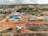 An aerial view captures a construction site with machinery and materials scattered across a reddish dirt ground. Several buildings and storage units are positioned at the site, along with a small number of vehicles. Surroundings include residential structures, green areas with palm trees, and hilly terrain in the background under a cloudy sky.