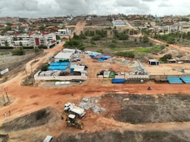 An aerial view captures a construction site with machinery and materials scattered across a reddish dirt ground. Several buildings and storage units are positioned at the site, along with a small number of vehicles. Surroundings include residential structures, green areas with palm trees, and hilly terrain in the background under a cloudy sky.