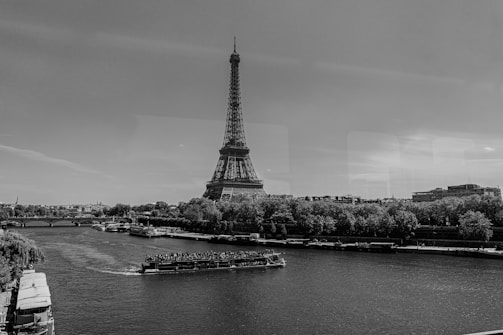 A black and white photograph features the Eiffel Tower in the background, positioned next to the River Seine. A boat filled with passengers is seen cruising along the river. Trees and buildings line the riverbank, creating a blend of natural and urban elements under a clear sky.