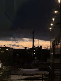 An industrial facility is silhouetted against the backdrop of a dramatic sunset. The sky transitions from a warm orange near the horizon to dark blue higher up. Streetlights illuminate the foreground, casting a subtle glow on the building to the right. Snow covers parts of the ground, adding to the evening's cold atmosphere.
