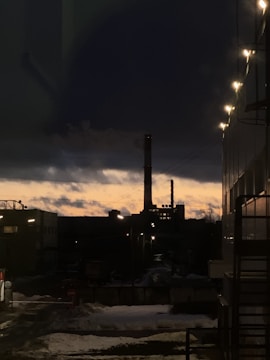 An industrial facility is silhouetted against the backdrop of a dramatic sunset. The sky transitions from a warm orange near the horizon to dark blue higher up. Streetlights illuminate the foreground, casting a subtle glow on the building to the right. Snow covers parts of the ground, adding to the evening's cold atmosphere.