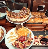 A variety of cakes and pastries are arranged on plates. One cake is topped with chocolate, cream, and garnished with orange slices and pomegranate seeds. There is a partially eaten cake with fruit toppings, including cherries and citrus slices. Almond-decorated pastries are also visible, neatly arranged on a tray.