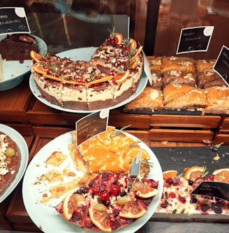 A variety of cakes and pastries are arranged on plates. One cake is topped with chocolate, cream, and garnished with orange slices and pomegranate seeds. There is a partially eaten cake with fruit toppings, including cherries and citrus slices. Almond-decorated pastries are also visible, neatly arranged on a tray.