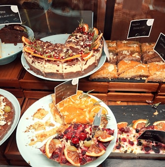 A variety of cakes and pastries are arranged on plates. One cake is topped with chocolate, cream, and garnished with orange slices and pomegranate seeds. There is a partially eaten cake with fruit toppings, including cherries and citrus slices. Almond-decorated pastries are also visible, neatly arranged on a tray.