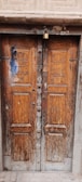 A rustic wooden double door with intricate carvings and a large lock at the top. The wood shows signs of aging and weathering, with minor cracks and discoloration. The door stands within a stone or brick frame, giving a sense of history and wear.