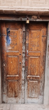 A rustic wooden double door with intricate carvings and a large lock at the top. The wood shows signs of aging and weathering, with minor cracks and discoloration. The door stands within a stone or brick frame, giving a sense of history and wear.