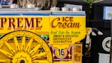 A colorful ice cream cart is adorned with vibrant text and decorative designs. The cart has a striking yellow color with red and blue accents, featuring large wheels and stainless steel lids on top. The words 'Supreme Ice Cream' and various contact numbers are prominently displayed, along with the phrase 'Accept Order Any Occasion'. The phrase 'In God We Trust' is also visible on the cart.