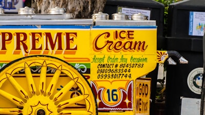A colorful ice cream cart is adorned with vibrant text and decorative designs. The cart has a striking yellow color with red and blue accents, featuring large wheels and stainless steel lids on top. The words 'Supreme Ice Cream' and various contact numbers are prominently displayed, along with the phrase 'Accept Order Any Occasion'. The phrase 'In God We Trust' is also visible on the cart.