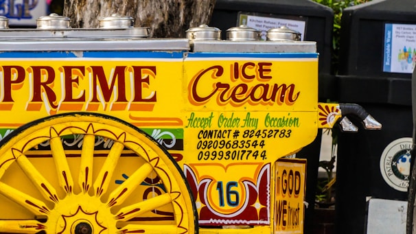 A colorful ice cream cart is adorned with vibrant text and decorative designs. The cart has a striking yellow color with red and blue accents, featuring large wheels and stainless steel lids on top. The words 'Supreme Ice Cream' and various contact numbers are prominently displayed, along with the phrase 'Accept Order Any Occasion'. The phrase 'In God We Trust' is also visible on the cart.
