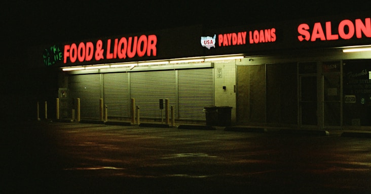 A dimly lit strip mall at night with businesses including food and liquor, payday loans, and a salon, highlighted by neon red signage reflected on wet pavement.