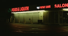 A dimly lit strip mall at night with businesses including food and liquor, payday loans, and a salon, highlighted by neon red signage reflected on wet pavement.