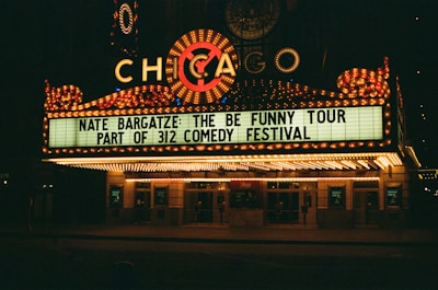 A brightly lit theater marquee displays information about Nate Bargatze's comedy tour, part of the 312 Comedy Festival. The marquee is adorned with numerous lights and features the word 'Chicago' prominently.
