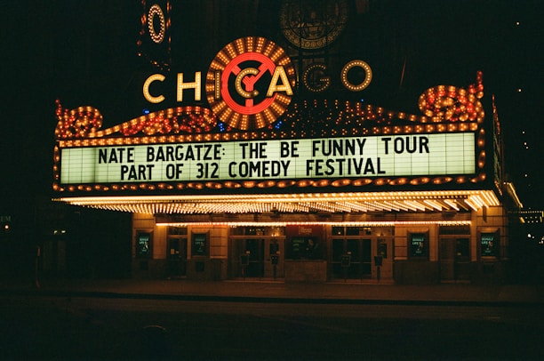 A brightly lit theater marquee displays information about Nate Bargatze's comedy tour, part of the 312 Comedy Festival. The marquee is adorned with numerous lights and features the word 'Chicago' prominently.
