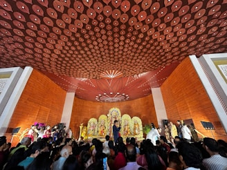 A large crowd gathers in a hall with ornate decorations to observe a religious ceremony. The ceiling features intricate patterns, and the space is lined with orange wooden panels. At the front, there is an elaborate altar displaying brightly adorned deities with floral arrangements. People are standing and some are taking photos, creating an atmosphere of reverence and celebration.
