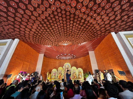 A large crowd gathers in a hall with ornate decorations to observe a religious ceremony. The ceiling features intricate patterns, and the space is lined with orange wooden panels. At the front, there is an elaborate altar displaying brightly adorned deities with floral arrangements. People are standing and some are taking photos, creating an atmosphere of reverence and celebration.