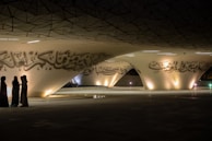 A modern architectural space featuring large, sweeping curves with Arabic calligraphy adorning the surfaces. Three people are standing together in discussion, dressed in traditional Middle Eastern attire. The area is dimly lit with soft, warm lights highlighting the calligraphic details.
